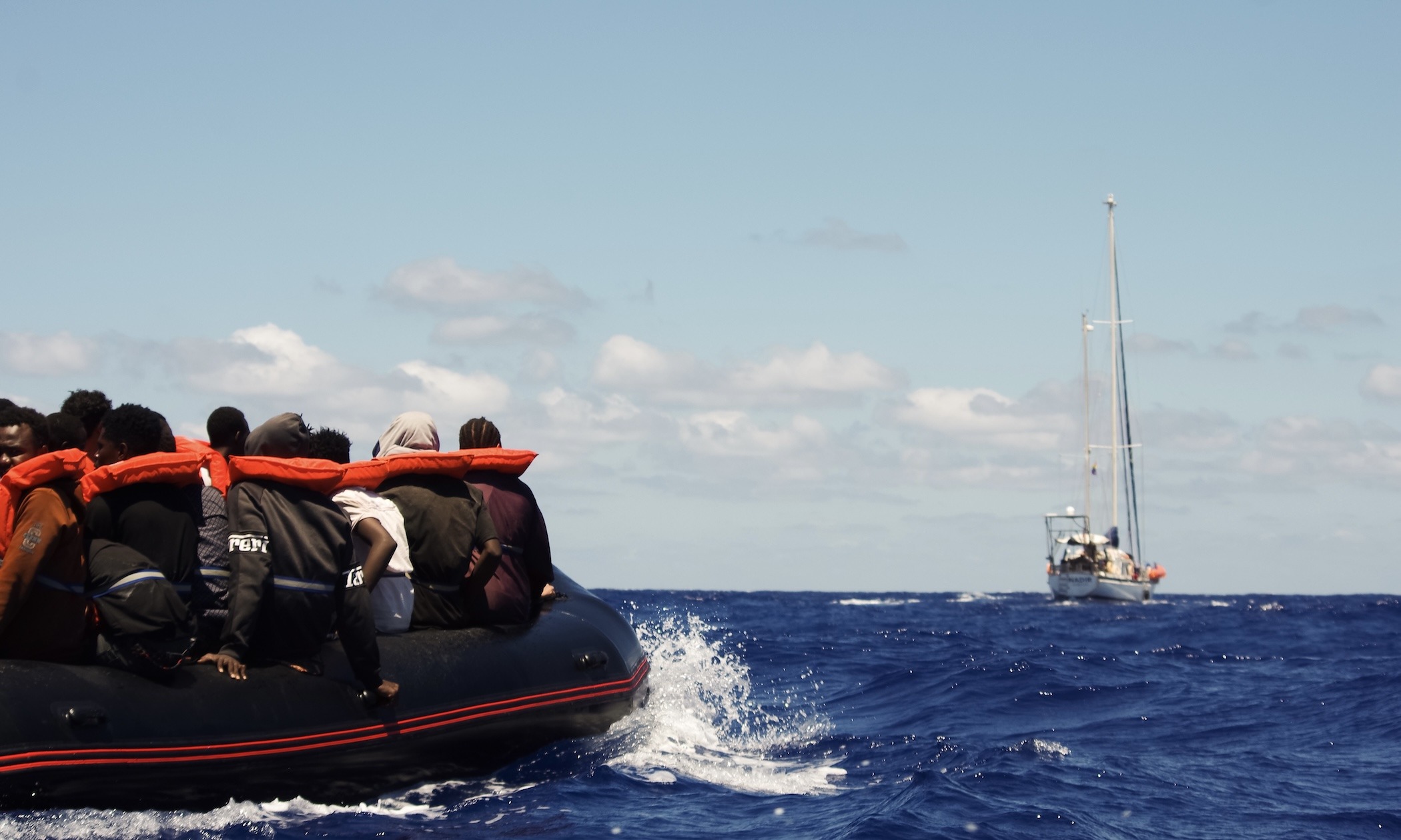 Die Überlebenden auf dem Schlauchboot nachdem die Crew der NADIR Rettungswesten und Trinkwasser verteilt hat. Im Hintergrund zu sehen: Das Segelschiff NADIR, das Such- und Rettungseinsätze fährt. 