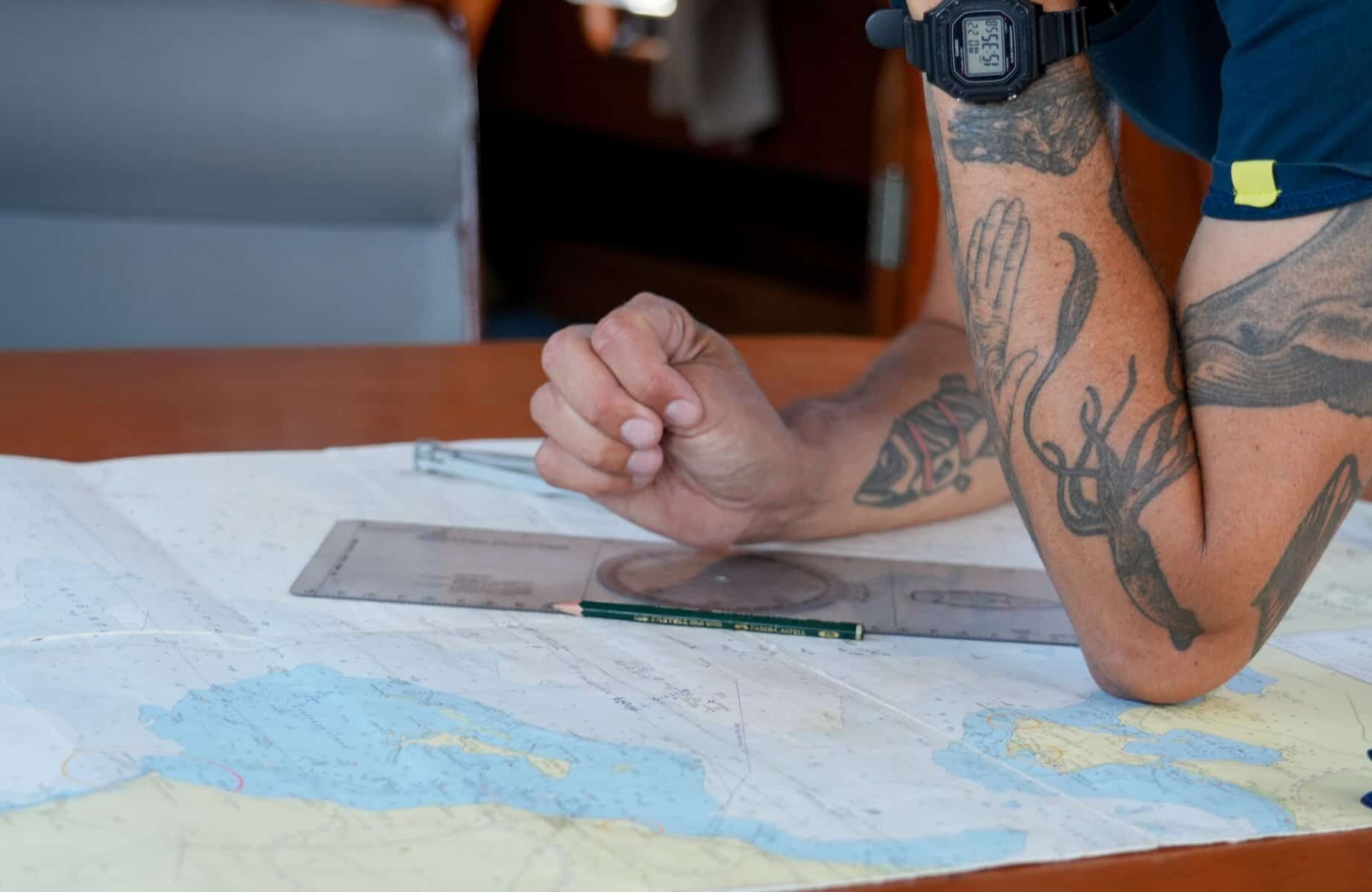 Person mit Tattoos prüft Karte und Dokumente auf einem Tisch, Fokus auf Hand und Unterarm.