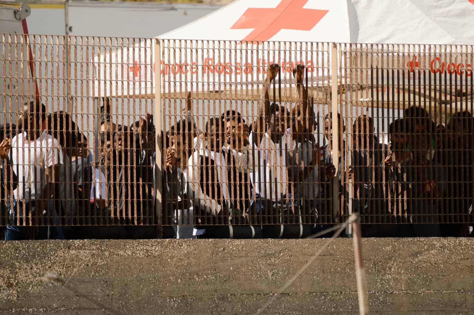 Gruppe von Menschen steht hinter einem Metallzaun vor einem Zelt mit dem Symbol des Roten Kreuzes.