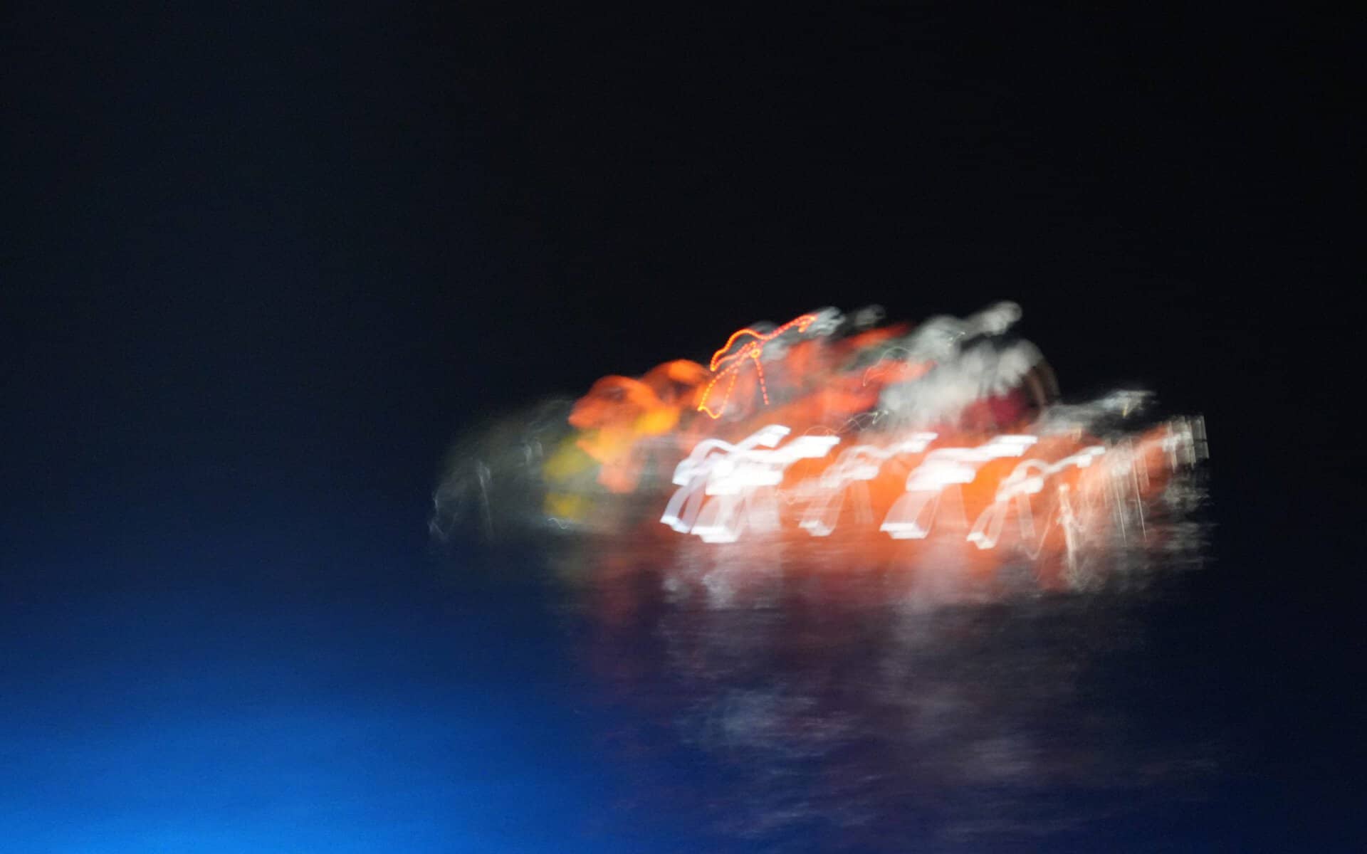 Unscharfe Aufnahme eines orangefarben beleuchteten Schlauchboots mit mehreren Personen auf der Flucht auf dunklem Meer bei Nacht.